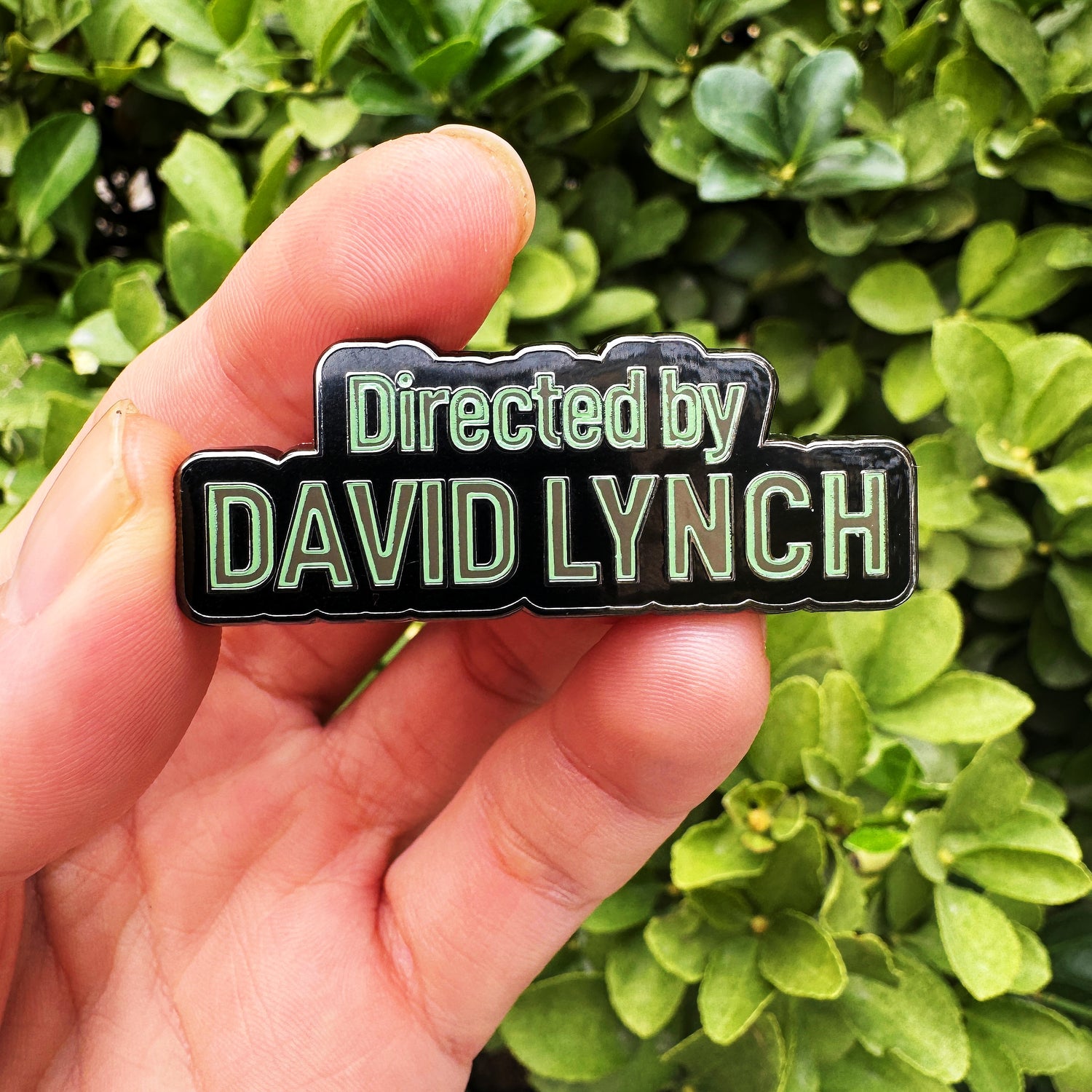 Hand holding a 'Directed by David Lynch' enamel pin against a green leafy background